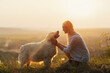 Happy young woman is sitting on the hill at sunset lovingly hugging her large breed dog.