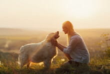 Happy Young Woman Is Sitting On The Hill At Sunset Lovingly Hugging Her Large Breed Dog.