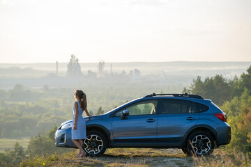 Wall Mural - Happy young woman driver in blue dress enjoying warm summer evening standing beside her car. Travelling and vacation concept