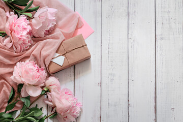 fresh pink peony flowers and gift in craft paper on white wooden background, space for text