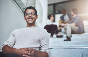 Poster - Weve taken some exciting steps as a company. Portrait of a young designer sitting on a staircase with his colleagues in the background.