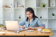 © amnaj - Charming Asian woman with a smile sitting and taking notes with computer laptop on her desk, enjoying work.