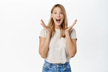 Wall Mural - Surprised and excited blond girl screams of joy, reacting amazed and happy at promo sale, super big news, standing over white background