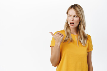 Wall Mural - Annoyed girl pointing and looking left with disdain, upset by something, complaining, standing in t-shirt over white background