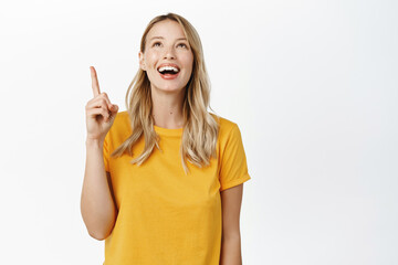 Wall Mural - Excited smiling blond girl looking and pointing up with amused face, reading promo text, banner on top, standing in yellow tshirt over white background