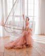 young woman with short black hair in pink princess dress with train is sitting on the crystal chandelier near huge panoramic window background like dior style. fashion concept, free space