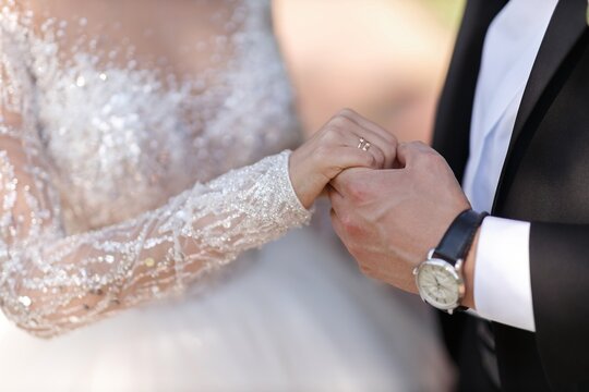 a man in a black tuxedo and a white shirt with a classic watch gently holds the hand of the bride in a white luxurious dress