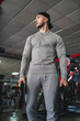 Chico joven musculoso con chándal gris y gorra negra posando en un gimnasio rodeado de maquinas de musculación 