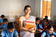 Smiling Teacher with books in hand standing at classroom in between students at classroom - concept of professional occupation, woman empowerment and education