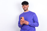 Fototapeta  - young arab man with curly hair wearing purple sweatshirt over white background Mock up copy space. Using mobile phone, typing sms message