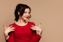 Young Woman In Red Dress Points To Herself With Hands. Elegant Girl Posing In Studio On Beige Background. Self-love. Treat Yourself. Workspace Area