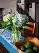 A clear glass vase of pale blue and white hydrangea's sit on a dining room table next to a wooden bowl of green avocados. Soft focus view of homes living room in the backgrounds.