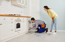 Plumber Crouching On The Floor In A Modern White Kitchen And Using A Drain Cable To Clean A Clogged Sink Pipe. Young Girl Calls The Plumber To Fix The Problem Of A Clogged Drain Under Her Kitchen Sink