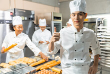 Smiling Young Man Bakers Looking At Camera..Chef  Baker In A Chef Dress And Hat, Cooking Together In Kitchen.Young Asian Man Takes Fresh Baked Cookies Out Of Modern Electric Oven In Kitchen.
