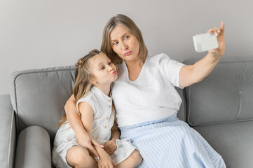 A little girl and her grandmother take selfies on the phone and curve her lips with a duck. Cozy home sofa.