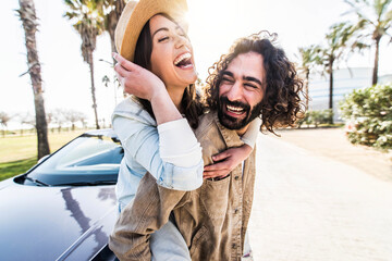 Playful couple of lovers having piggyback ride near the car - Happy tourists enjoying road trip on summertime day out - Vacations, youth lifestyle and transportation concept