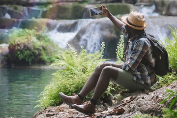 Wall Mural - African Man Traveller with backpack sitting for selfie and relaxing freedom at the waterfall.