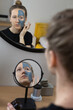 © Богдан Перфильев - rear view photo of a young caucasian woman looking at herself in small mirror, applying mud mask other face, reflection of beauty treatment in the big round mirror on the wall at home