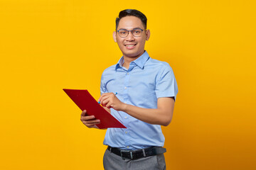 Portrait of handsome young Asian businessman in glasses standing confidently holding document folder with smiling face isolated over yellow background