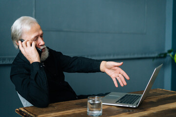 Wall Mural - Front view of angry gray-haired aged male talking to tech support and asking for help repairing broken laptop computer sitting at wooden table, at home office.