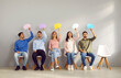 © Studio Romantic - Group of people sitting on row of chairs, thinking about something and holding empty colorful mock up speech bubbles. Young male and female company employees taking part in survey and sharing opinions