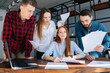 © dikushin - Front view of tired young female team leader under stress at office sitting at desk with multi-ethnic colleagues shaking paper documents standing near her. Woman boss exhausted by management work.