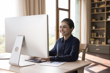 Canvas Print - Smiling Indian female freelancer work on desktop pc at home office enjoy creative job. Glad young woman manager look at computer screen analyze statistic information satisfied with good sales result