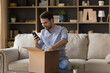 Excited guy blogger sit on couch unboxing postal package with consumer goods received from web shop by mail shoot video on mobile phone webcam. Happy young male open parcel delivery make photo on cell