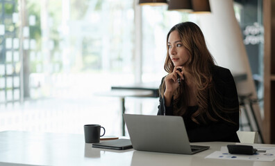 Portrait of asian businesswoman sitting with laptop computer looking outside while working with paperwork in the office