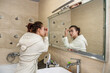 © RomanR - beautiful brunette near the mirror applies foundation on her face for makeup.