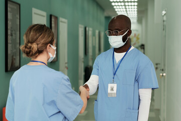 Aufkleber - Young African American surgeon in uniform and protective mask shaking hand of his female assistant while both standing in hospital corridor