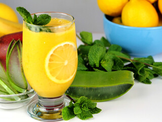 A drink made from banana, mango, lemon juice and fresh Aloe Vera. A glass with a yellow smoothie and a slice of lemon inside. Fresh fruits, Aloe Vera leaf pieces, mint leaves. On a white background