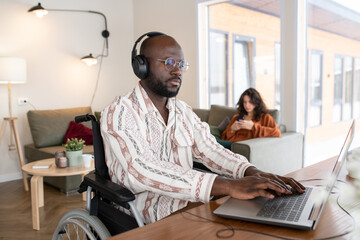 Aufkleber - Serious African American businessman in headphones typing by table in living room while working with online data against young woman