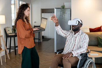 Aufkleber - Young woman with laptop standing in front of man with disability sitting in wheelchair with vr headset and pointing at virtual screen