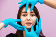 © _KUBE_ - Portrait of a mature pretty woman with cosmetologists' hands in rubber gloves at her face. Lilac background. The concept of plastic surgery and rejuvenation