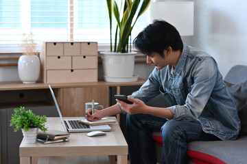 Wall Mural - Young man in casual clothes holding mobile phone and working or surfing internet with laptop computer.