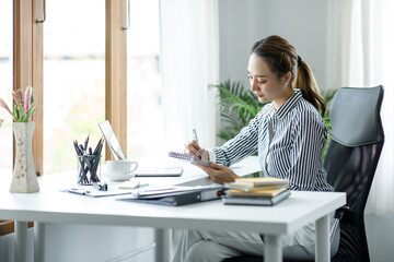 Smiling young Asian businesswoman working on a laptop computer at her desk in a bright modern home office, doing calculating expense financial report finance making notes on paper graph data document.