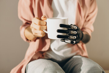 Artificial bionic female hand with mechanical metal fingers holding cup of hot drink in white cup. Woman training new skills after surgery of arm amputation. Cyborg people. Cybernetics, innovations