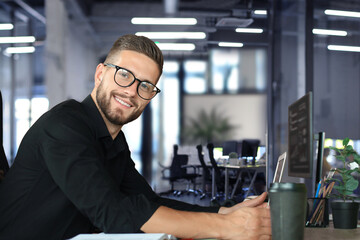 Wall Mural - Young business man is using laptop while sitting in the office, looking at camera, smiling