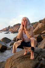 Woman In Squatting Position With Eyes Closed Touching Cheek On Rock At Beach