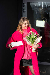 Vertical portrait of glad happy laughing blond woman in pink outfit, presents, bouquet tulips. Walking corporate party.