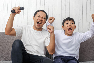 Excited expressions of father and son watching television  to their favorite football team making a goal and winning,  wearing white t shirt suitable for apparel mock up 