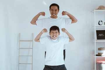 Asian young father in white t shirt and his son in white t shirt are smiling while pose showing muscle, spending time together at home