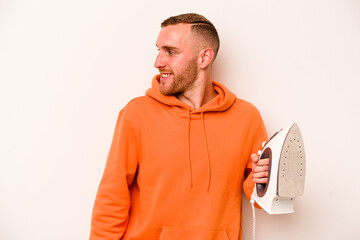 Young caucasian man holding iron isolated on white background looks aside smiling, cheerful and pleasant.