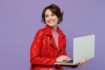 Wall Mural - Young smiling satisfied fun woman 20s in red leather jacket hold use work on laptop pc computer look aside on workspace area mock up isolated on plain pastel light purple background studio portrait.
