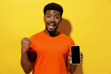 Young Man Of African American Ethnicity 20s In Orange T-shirt Hold In Hand Use Mobile Cell Phone With Blank Screen Workspace Area Do Winner Gesture Isolated On Plain Yellow Background Studio Portrait