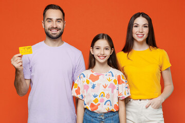 Wall Mural - Young smiling parents mom dad with child kid daughter teen girl in basic t-shirts hold in hand credit bank card isolated on yellow background studio portrait. Family day parenthood childhood concept