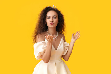 Wall Mural - Beautiful young African-American woman in stylish dress and tiara blowing kiss on yellow background