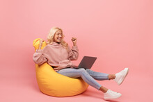 Overjoyed Woman Using Laptop And Raising Hands, Reading Great News And Celebrating Success, Sitting In Beanbag Chair