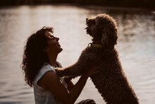 .Curly-haired Woman Playing With Her Brown Spanish Water Dog On A Lake's Floating Walkway. Silhouette